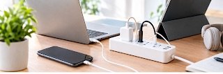 White power strip on a wooden desk with a laptop, smartphone, wireless headphones, and a green potted plant