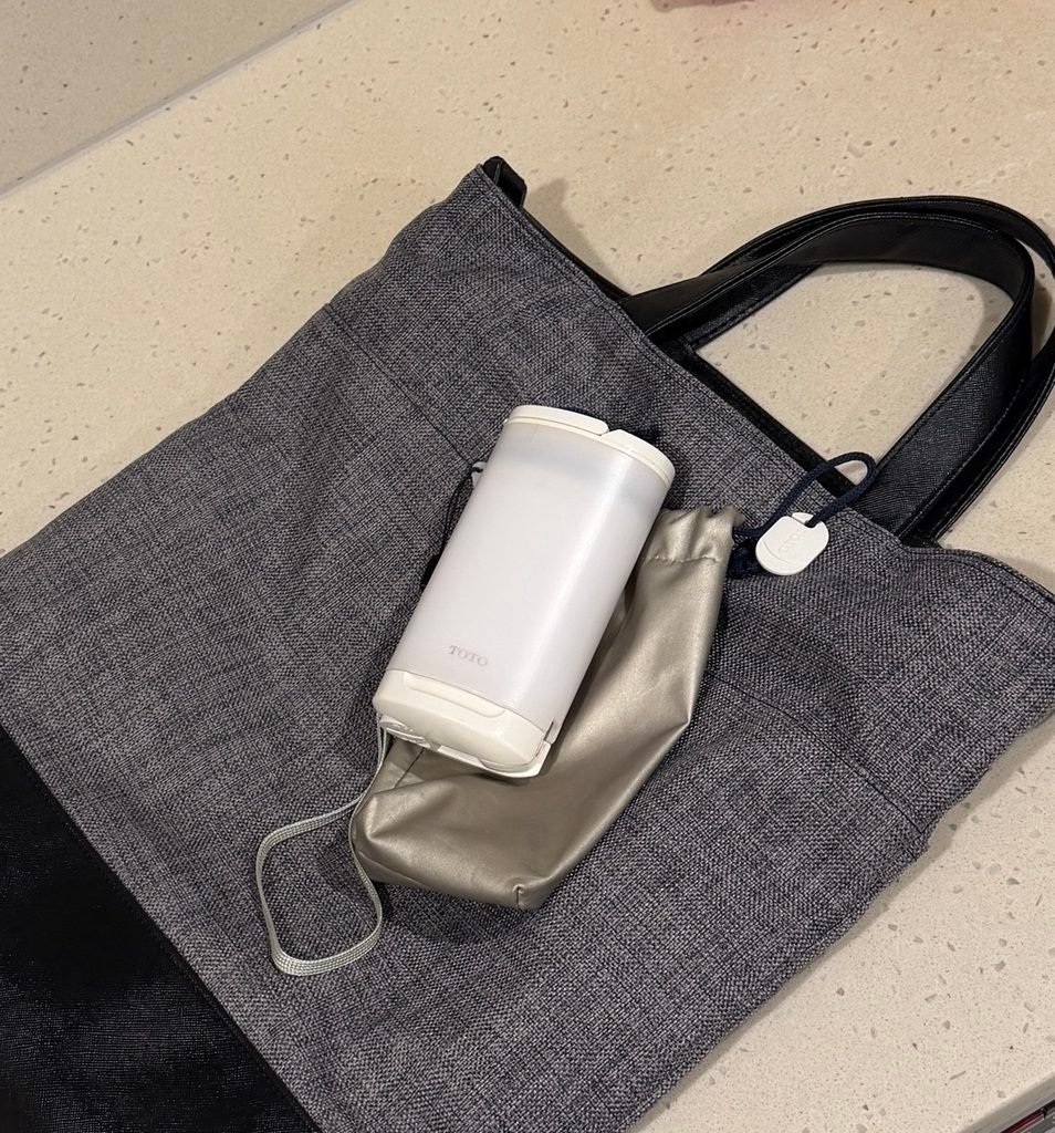 A TOTO travel washlet and a grey pouch sitting on a grey and black tote bag.