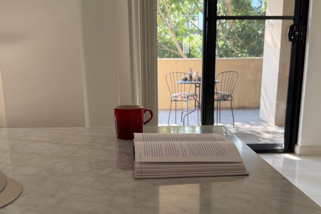 A red mug and an open paperback book on a white marble table next to a glass sliding door to a patio.